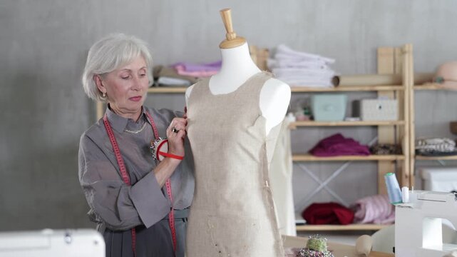 Mature dressmaker with a measuring tape stands in the sewing workshop near a mannequin and checks quality of the sewn sample, she looks at the stitches and makes corrections.