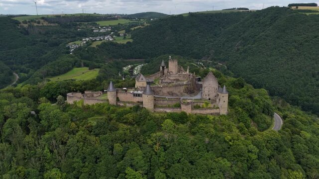 Majestic bourscheid castle sitting atop a forested hill with a winding road below and a lush valley stretching into the distance, showcasing a famous historical landmark in luxembourg
