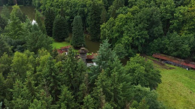 Beautiful aerial footage flying over a dense green forest, gradually revealing a secluded, picturesque castle like house surrounded by a tranquil moat reflecting the surrounding trees