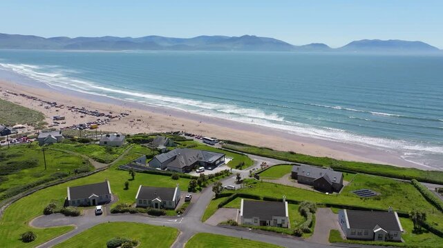 Rows of houses with manicured lawns overlook the expansive Inch Beach. Waves gently lap the sandy shore, with distant mountains framing the horizon. Perfect for travel and real estate content.