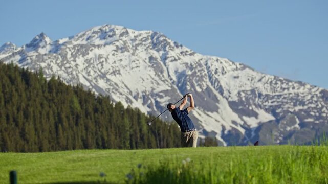 Professional golfer taking a full swing on the fairway at Verbier Golf Club in Switzerland. The snow-covered peak of Le Rogneux rises above the alpine course, creating a spectacular mountain golf sett