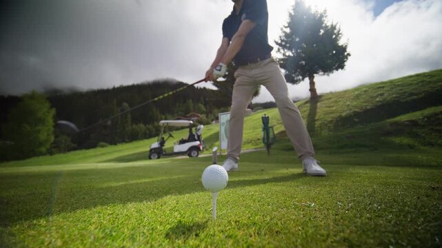Professional golfer striking a driver tee shot at Verbier Golf Club in Switzerland. Low-angle view captures the moment of impact with the golf ball on the tee against a spectacular Swiss Alps backdrop
