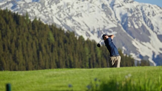 Professional golfer taking a full swing on the fairway at Verbier Golf Club in Switzerland. The snow-covered peak of Le Rogneux rises above the alpine course, creating a spectacular mountain golf sett