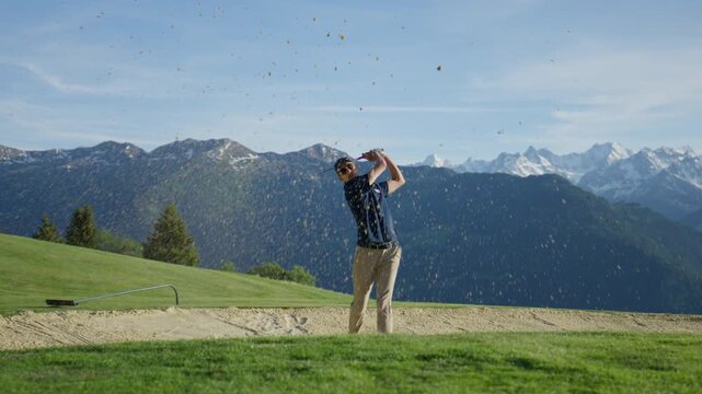 Professional golfer playing a bunker shot at Verbier Golf Club in Switzerland. Sand explodes from the bunker as the player executes a precise recovery shot with spectacular alpine peaks and mountain s