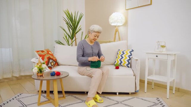 Focused talented elderly grandmother woman knitting sweater by yarn ball and needles with wrinkly hands crafting hobby in free time sitting comfortably on sofa. People, age and leisure.