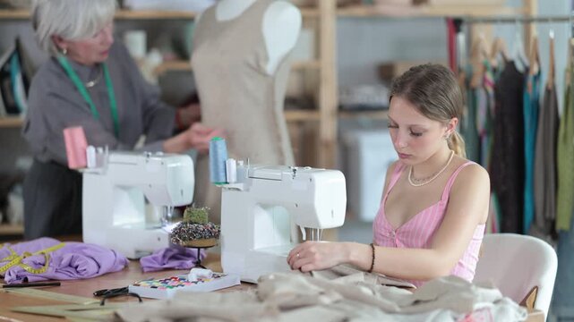 Young dressmaker works on a sewing machine against the background of a mature worker who checks a sample of a dress on a mannequin. Dressmakers work in the sewing industry. High quality 4k footage