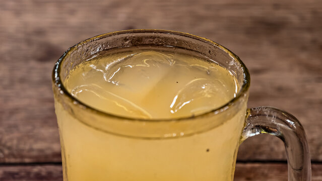 A refreshing cold drink of beras kencur in a clear glass, filled with ice cubes, on a rustic wooden surface