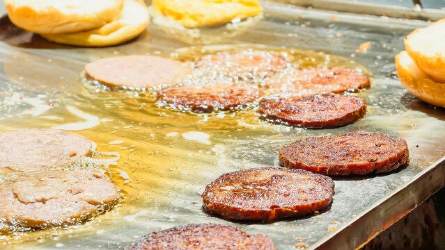 Juicy beef burger patties frying in oil on a flat top grill with buns