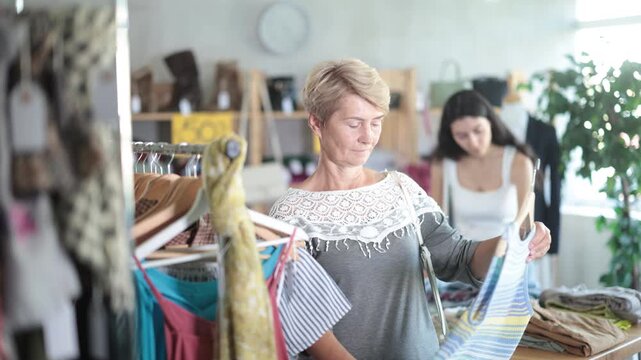 Senior woman customer making choice and elect something summer clothes in store. Modern urban fashion, casual practical clothes made of natural fabrics. Female client choosing something in background