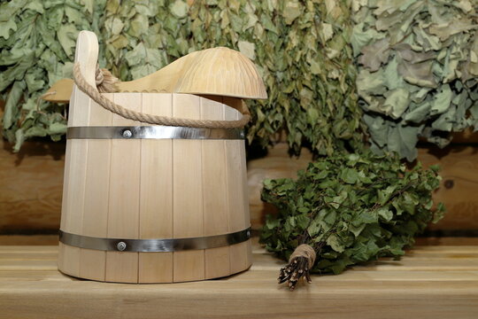 A wooden bucket with a wooden ladle and a birch broom stand on a plank bench against the background of a log wall with a set of dry birch and oak brooms is hanging on it.