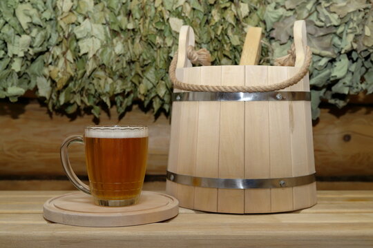 A mug of light beer on a circle wooden tray and a wooden bucket with a wooden ladle are on a plank bench against the background of a log wall with a set of dry birch and oak brooms is hanging on it.