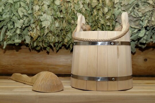 A wooden bucket and a wooden ladle stand on a plank bench against the background of a log wall with a set of dry birch and oak brooms is hanging on it. 