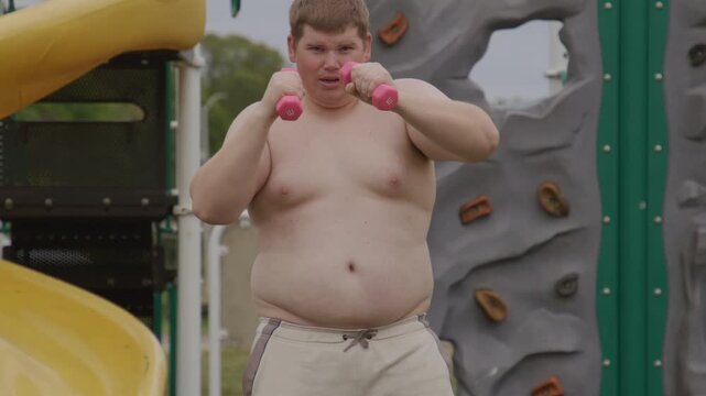 Overweight boy trains with dumbbells outdoors. Overweight guy does boxing exercise. Shirtless overweight boy punches with weights. Active overweight kid workout in park.