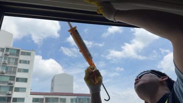 Professional maintenance worker wearing yellow protective gloves carefully applies silicone sealant using a caulking gun along the exterior window joint of a residential high-rise apartment building. 