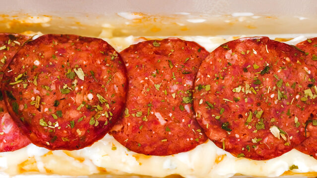 Close-up shot of seasoned pepperoni slices arranged in a row over creamy white sauce base