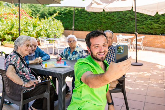 Happy young male caregiver taking a selfie with elderly residents enjoying a board game outdoors in a sunny retirement home