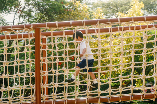 School-age boy climbing a rope bridge at an outdoor adventure playground in a sunny park. Concept of active childhood, physical development, balance skills, recreation, and healthy outdoor play.