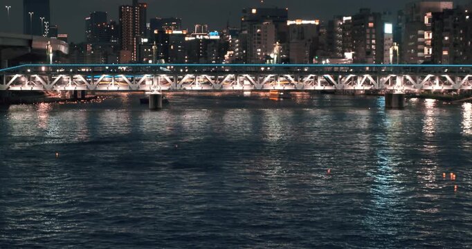 Illuminated railway bridge over dark river at night on cityscape backdrop, static wide view. No people. Panoramic bridge over sumida river reflecting in water waves in Tokyo darkness. Cityscape lights