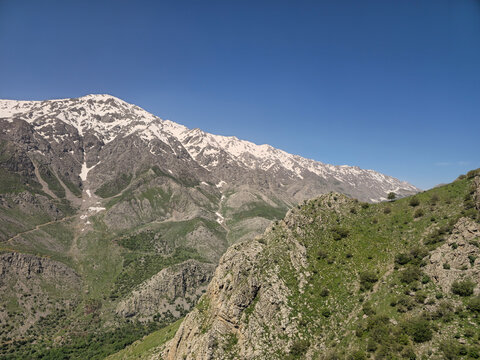 A green rocky ridge faces a massive snow-covered mountain range rising steeply above a deep spring valley under clear blue sky.
📍 Hawraman Takht, Kurdistan, Iran 