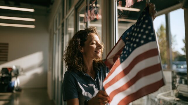 A smiling woman proudly decorates a modern office window with a small american flag to celebrate national pride in a bright professional workspace