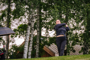 Fototapeta premium Female golfer in black athletic wear swings golf club on green course with trees and wooden structure in the background, cloudy sky overhead