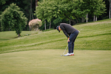 Obraz premium Male golfer in black long-sleeve shirt and navy pants prepares to putt on green golf course surrounded by trees and manicured grass in a sunny outdoor setting