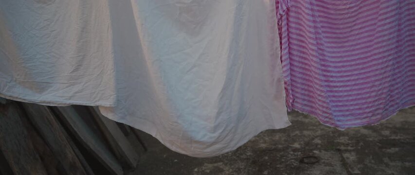 White and pink striped sheets hung on a clothesline outdoors, gently billowing over a rustic backyard surface. 4K.