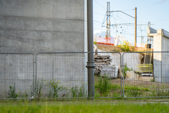 Industrial construction zone featuring a massive grey concrete bridge pillar behind temporary metal grid safety fencing alongside railway infrastructure