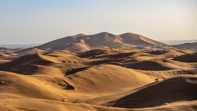 Sand dunes rise and fall in Erg Chebbi, near Merzouga in the Sahara Desert, under a clear sky. Visitors explore the vast sandy landscape during a warm day.