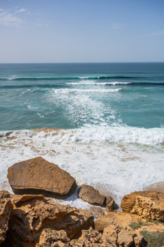 Waves crash on the rocky shore as the Atlantic Ocean meets the land in southern Morocco. The sky is clear and the water is a vibrant shade of blue under the sunlight.