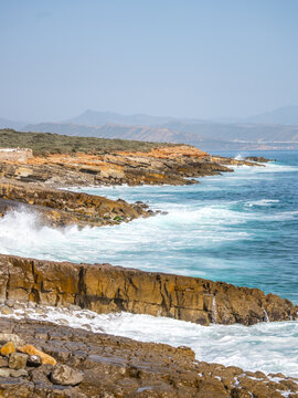 Waves crash against the rocky coast of the Atlantic Ocean in southern Morocco. The sun shines bright, and the landscape shows cliffs and water stretching towards the horizon.