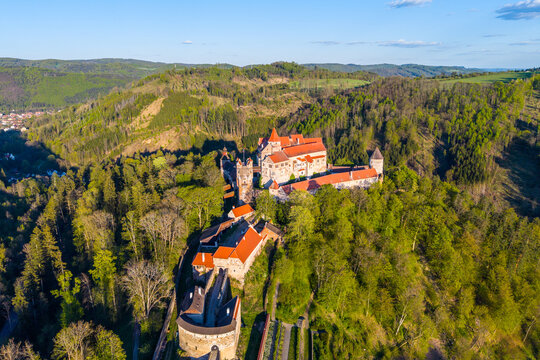 A view of Pernstejn castle from the sky shows its structure surrounded by trees and a hill. The landscape includes green forests and clear skies. This scene captures the medieval design.