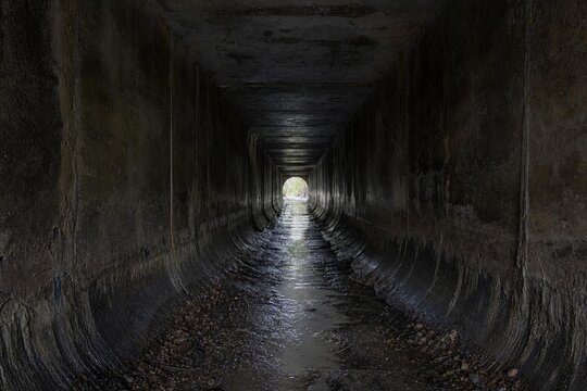 Inside a dark urban drainage culvert, wet walls leading to distant light.