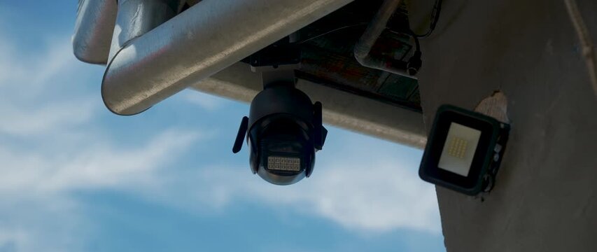 Outdoor security camera and surveillance system mounted under an eave, monitoring a building perimeter for safety and protection. Modern CCTV camera and floodlight. 4K.