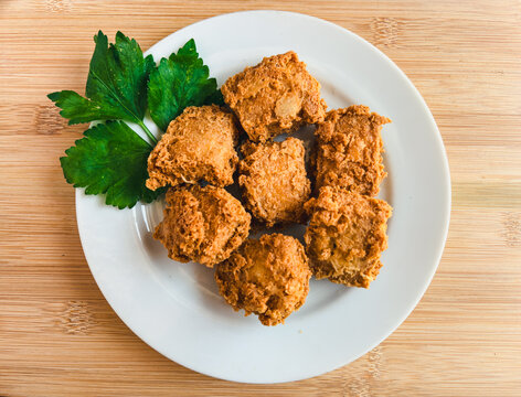 Crispy fried tofu or tahu crispy pieces sit on a white plate. Fresh parsley garnishes the dish, adding color. Wooden table provides a warm, rustic.  Simple, appetizing presentation draws the eye