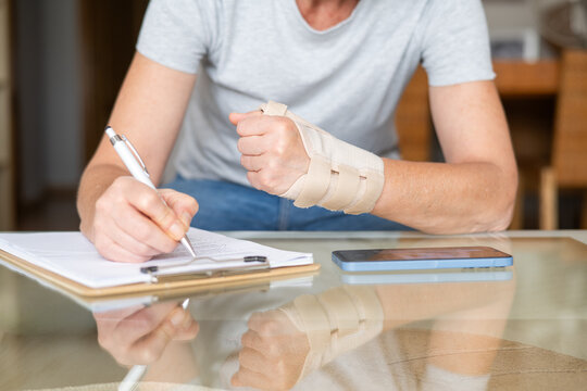 Woman with orthotic wrist splint fills out medical leave paperwork at desk, supporting painful hand while completing injury and disability forms for treatment and compensation
