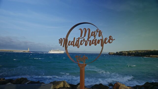 Mediterranean Sea sign in Tarifa, Spain, with a ferry crossing the Strait of Gibraltar near Isla de las Palomas. Iconic coastal landmark marking the meeting of the Mediterranean and Atlantic waters.