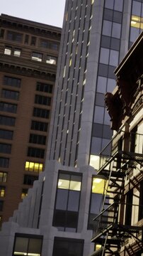 A mix of buildings in a city at night. A fire escape is visible on an older structure. Newer buildings with lights are seen in the background, showcasing urban architecture.