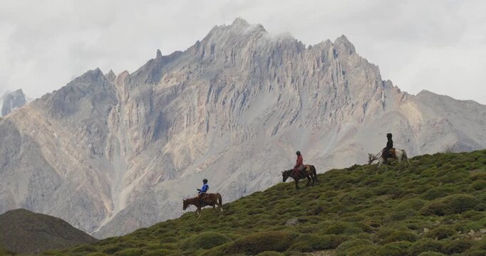 Aerial shot tracks across the green hills of Mustang, Nepal, moving in the opposite direction of horseback riders as they travel beneath a towering, rugged Himalayan peak