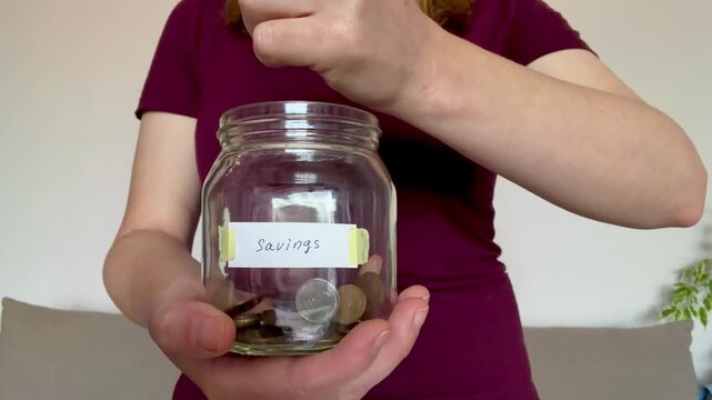 Woman putting coins into a glass jar labeled savings, money, finance, economy, investment, budget