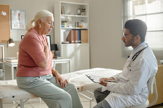 Senior woman consulting male doctor in clinic, describing chest pain during medical visit and receiving assessment. Useful for healthcare marketing, patient care, insurance