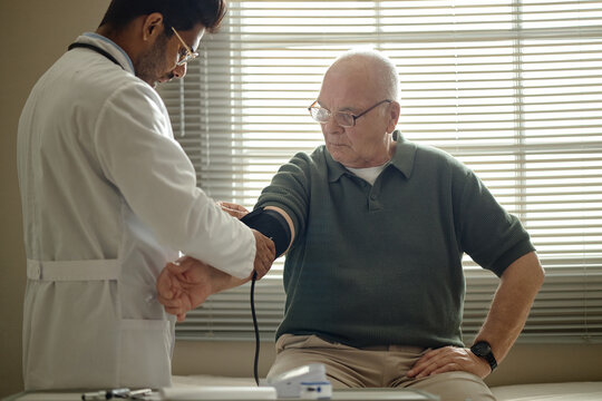 Senior man receiving blood pressure check from male doctor in clinic, monitoring cardiovascular health during medical visit. Ideal for healthcare, senior care, prevention campaigns