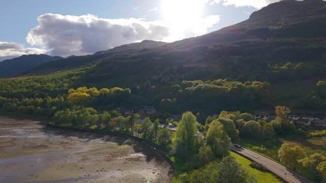 Drone rising over the head of Loch Long in the Scottish Highlands, showing the A83 road winding past the shoreline under beautiful early summer evening light.