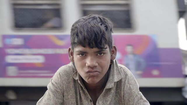 A daylight outdoor close up shot of a young Indian Asian untidy male kid or boy looking or facing the camera with a light smile, with a speedy local or suburban city train passing in the background