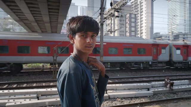 A daylight outdoor shot of a young Indian Asian poor male kid or boy next to railway tracks, looking or facing the camera with a serious or intense look on his face, speedy train passing in the back