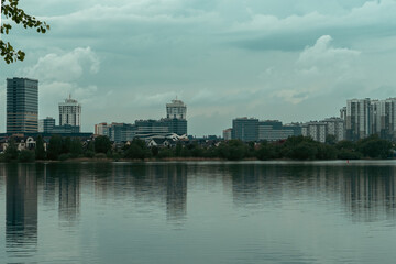 Fototapeta premium Modern Minsk residential district Lebyazhiy and business buildings reflected in calm water of Drozdy reservoir, urban waterfront landscape with green tree branches framing city skyline in Belarus