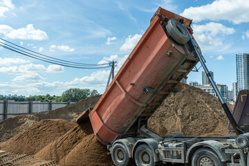 Dump truck unloading sand at urban construction site with high-rise buildings in background, heavy machinery working with bulk materials for city infrastructure development under blue sky © kalyanby