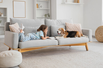Naklejka premium Cute little girl with Beagle dog reading book on sofa in living room