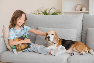 Naklejka premium Cute little girl with Beagle dog reading book on sofa in living room