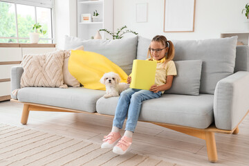 Naklejka premium Cute little happy girl in superhero costume with Maltese dog reading book on sofa at home